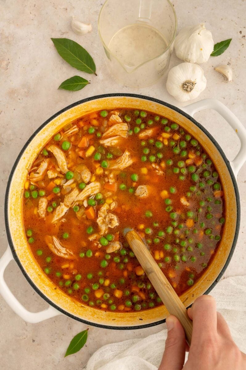 Peas and corn being stirred into a large pot of chicken barley soup with a wooden spoon.