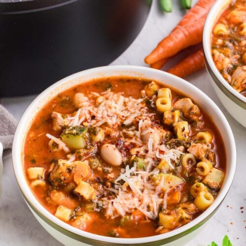 A bowl of chicken minestrone soup with carrots, celery, and a pot of the soup in the background.