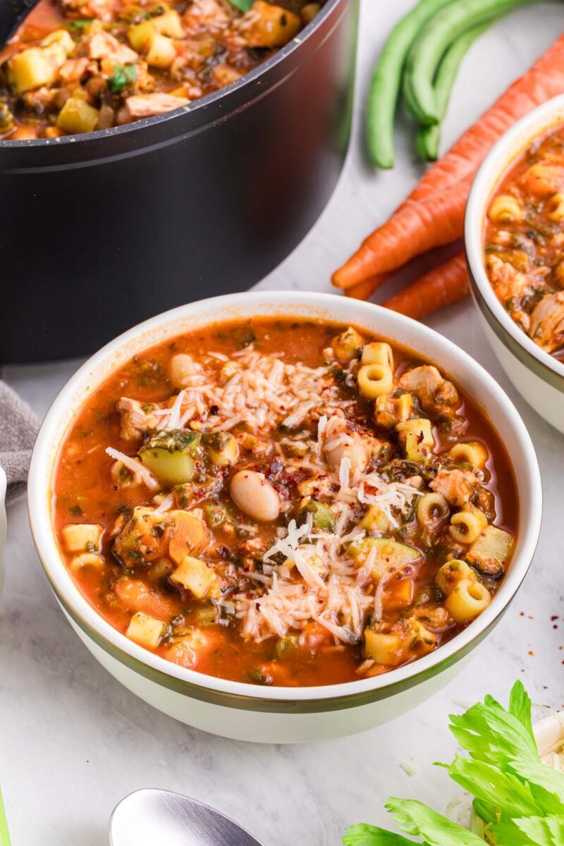 A bowl of chicken minestrone soup with carrots, celery, and a pot of the soup in the background.