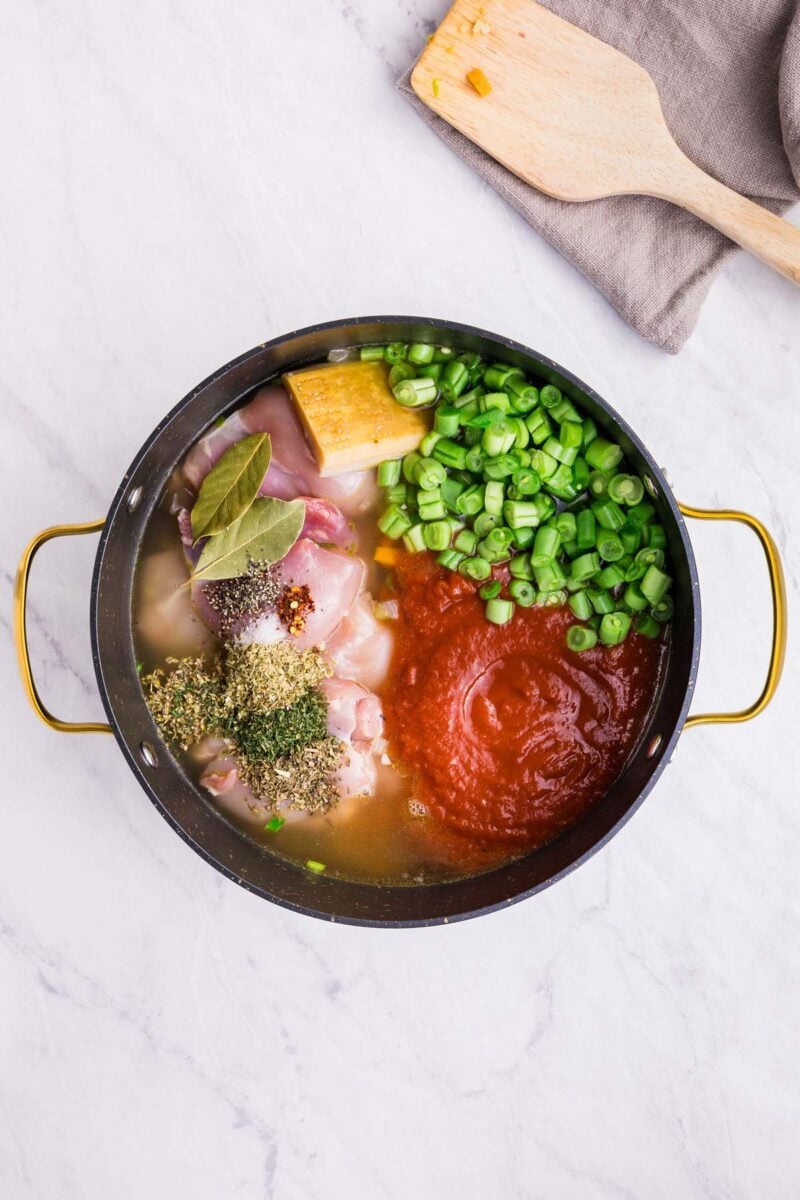Uncooked chicken thighs, chicken stock, tomatoes, water, green beans, spices, and parmesan cheese rind in a large pot to make chicken minestrone soup.