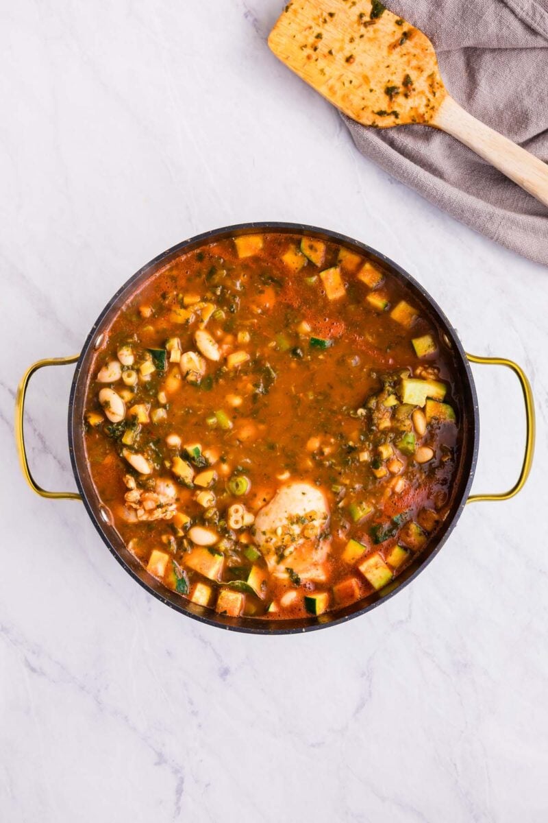 Cooked chicken minestrone soup in a stockpot before chicken is shredded with a spoon on the side.