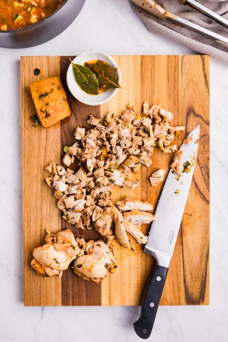 Chopped chicken on a wooden cutting board with a knife on the board and a discarded parmesan cheese rind and bay leaves in a bowl with a pot of chicken minestrone soup on the side.