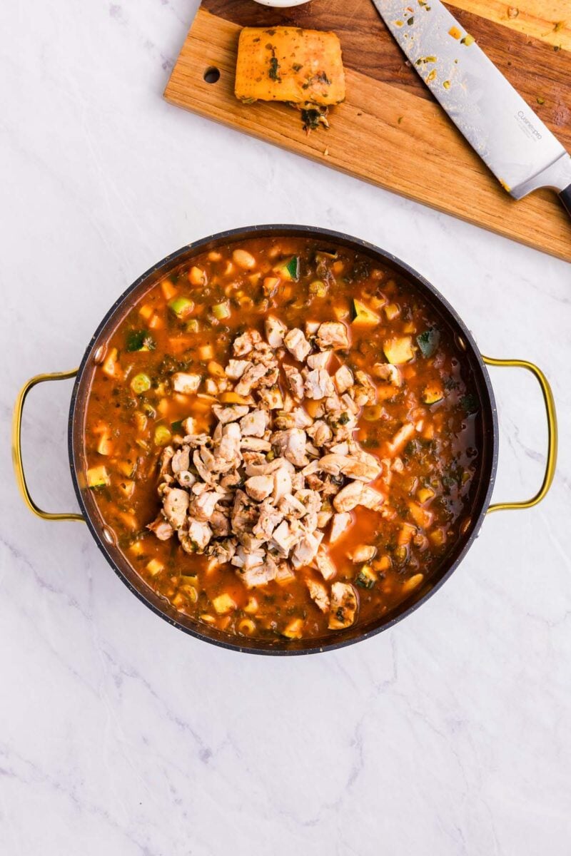 Chopped chicken added back to a large pot of chicken minestrone soup with a wooden cutting board and knife on the side.