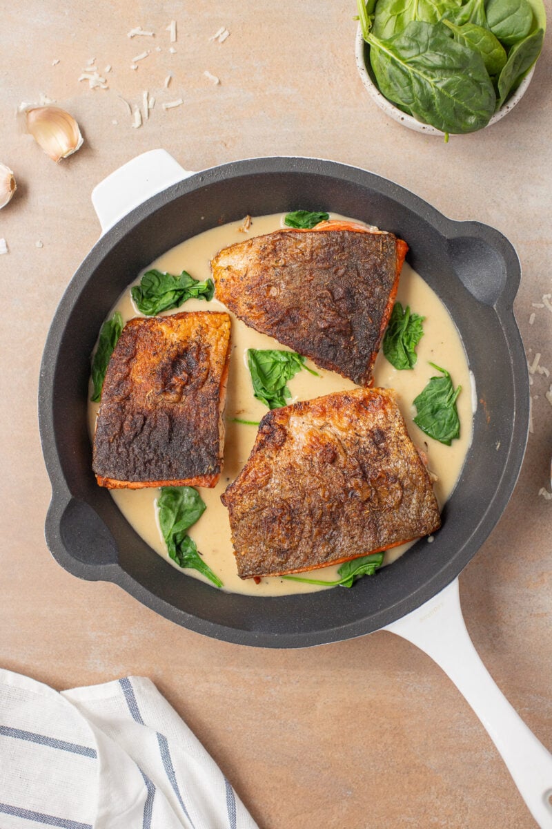 Three browned salmon filets in a pan with creamy garlic sauce with spinach in it.