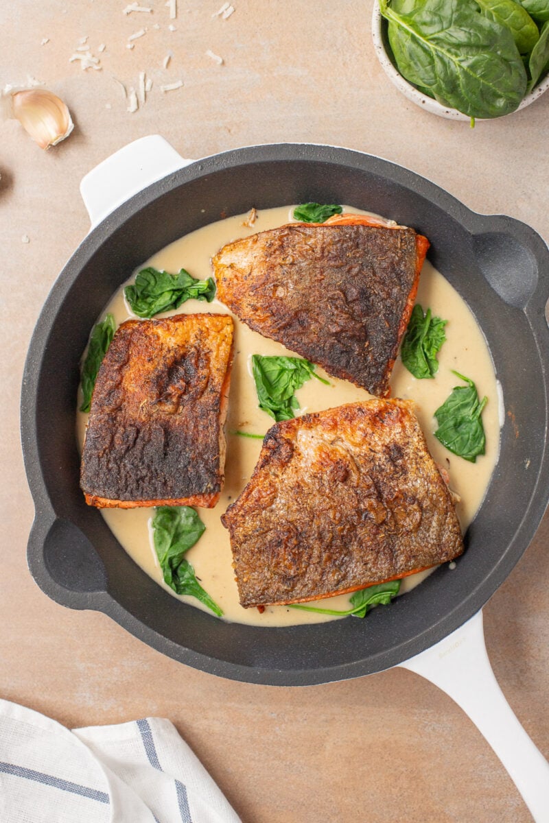 Three browned salmon filets in a pan with creamy garlic sauce with spinach in it.