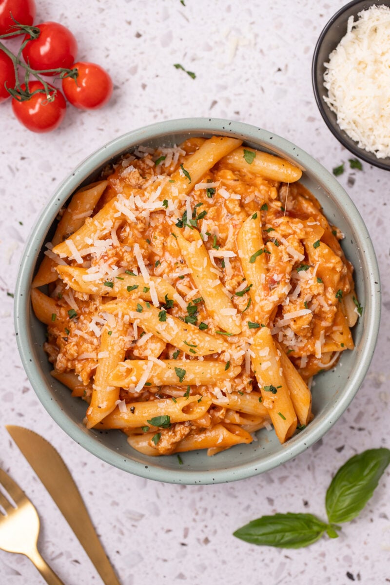 Ground Turkey Pasta in a bowl with tomatoes, cheese and herbs on the side.