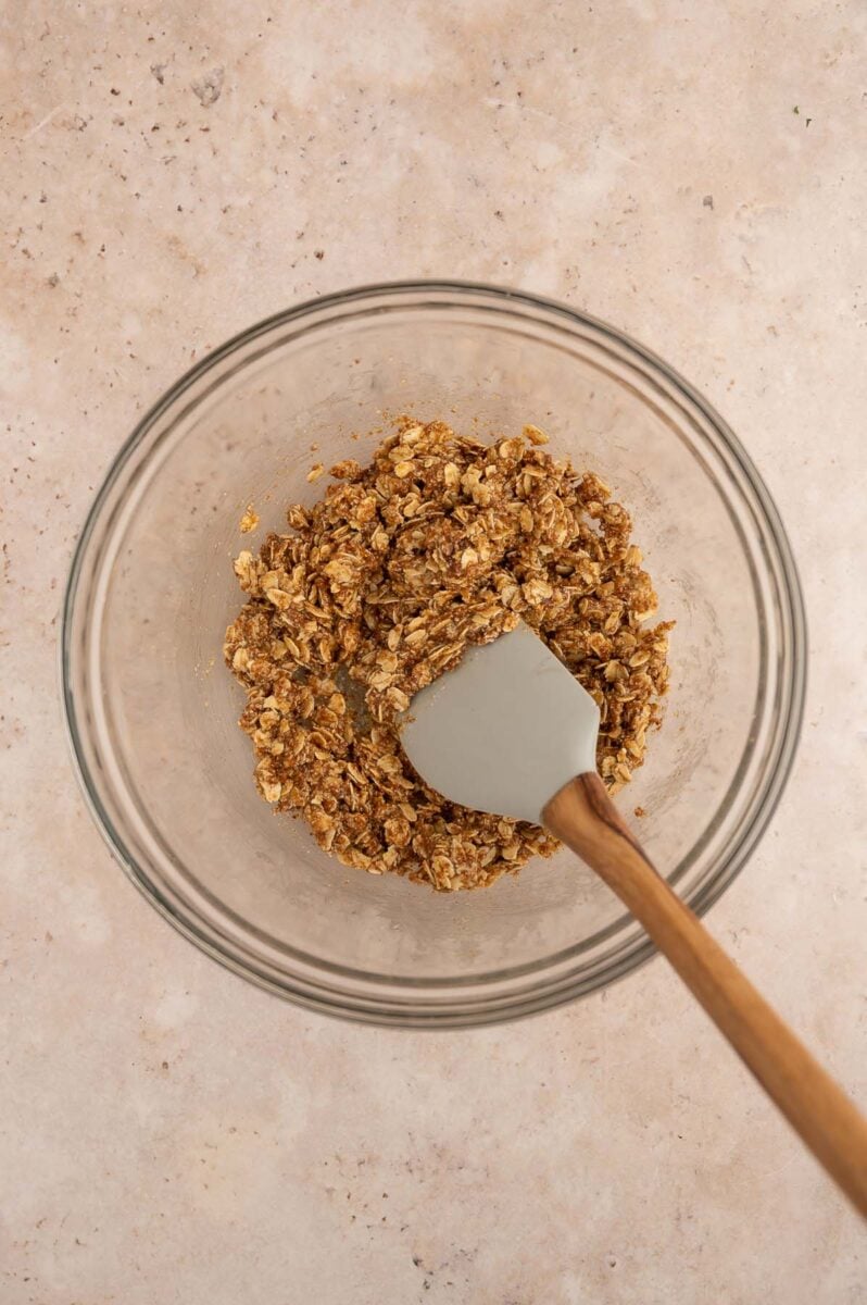 Apple Crisp topping ingredients mixed together in a clear glass bowl with a spatula resting in the bowl.