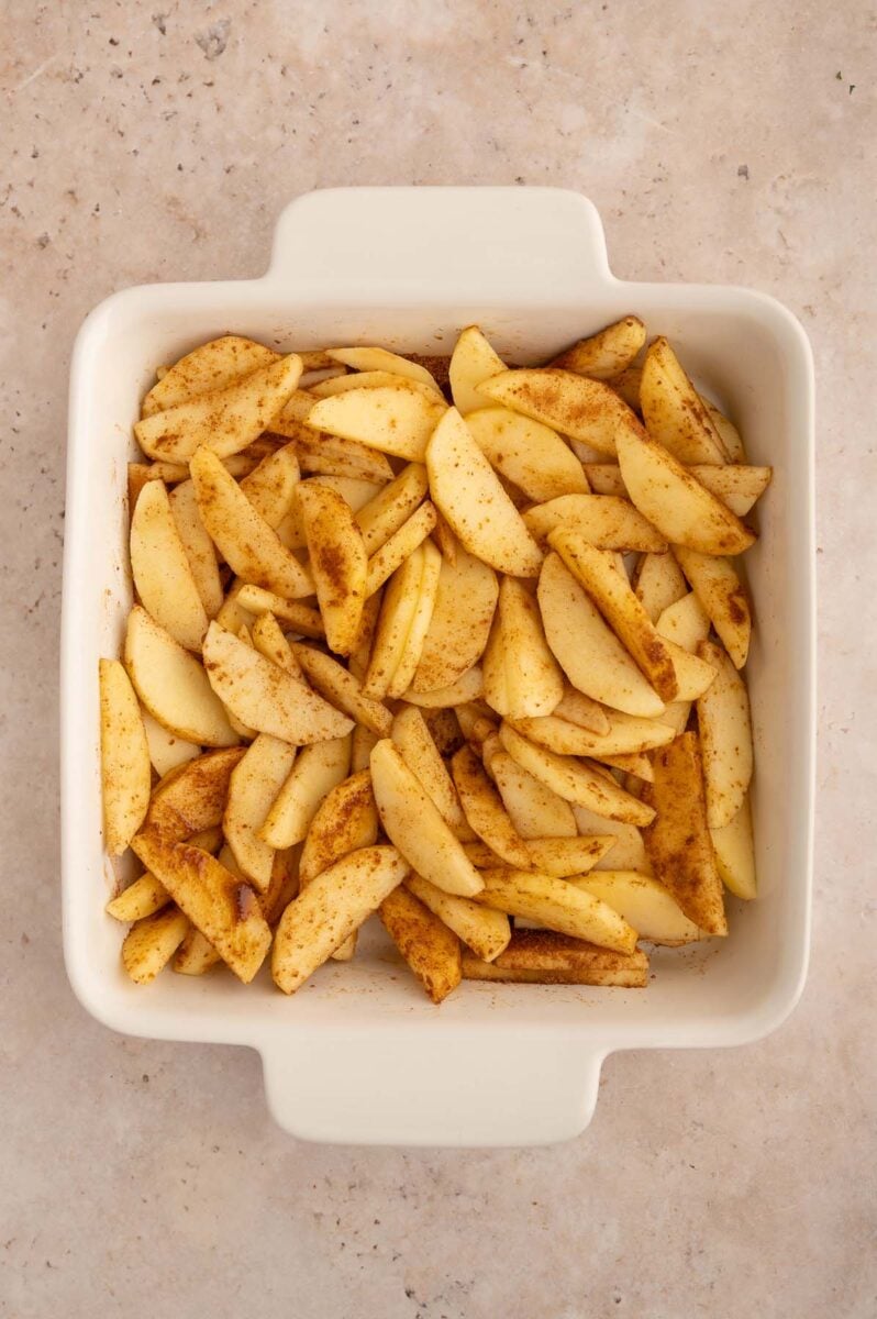 Sliced apples in a white baking dish tossed with spices before being baked.