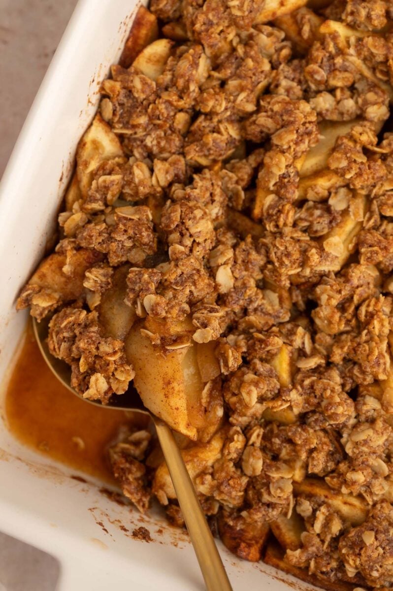 A close up of healthy apple crisp in a white baking dish with a gold spoon resting in it with a scoop taken out of the crips.