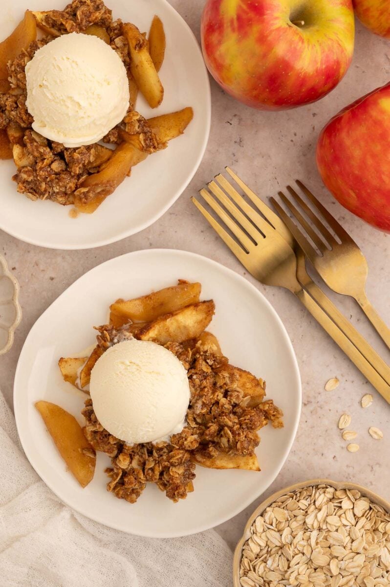Two white plates with a serving of healthy apple crisp on both topped with a scoop of vanilla ice cream with apples and forks on the side.