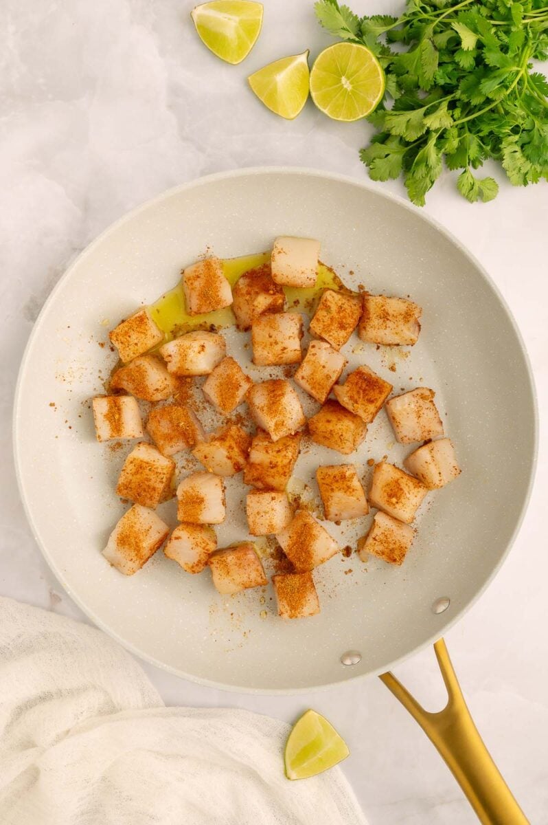 Uncooked seasoned fish pieces in a pan with fresh cilantro and limes on the side.