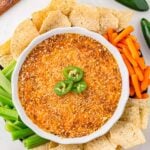 Healthy Jalapeno Popper Dip in a white round dish topped with jalapenos and surrounded by sliced celery, carrot sticks, and tortilla chips.
