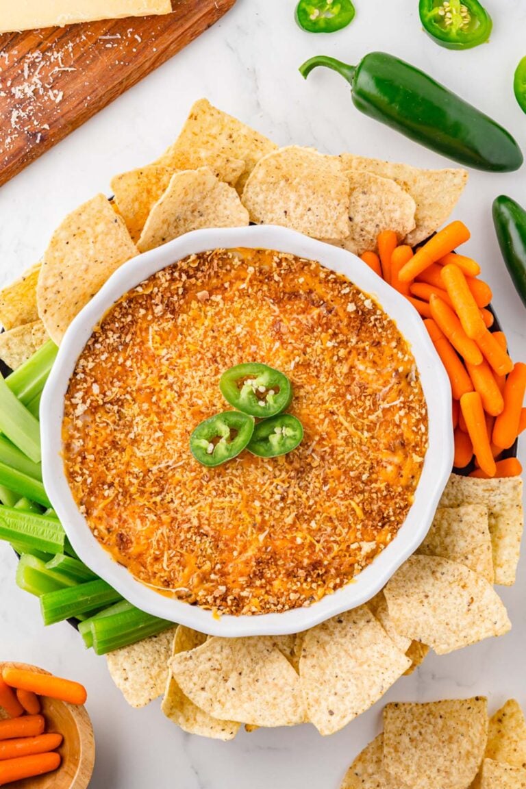 Healthy Jalapeno Popper Dip in a white round dish topped with jalapenos and surrounded by sliced celery, carrot sticks, and tortilla chips.