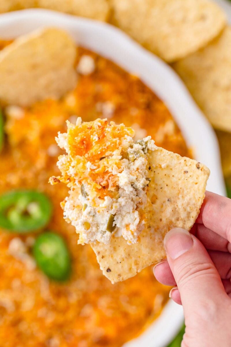 A hand holding up a tortilla chip with healthy jalapeno popper dip with the serving dish of the dip in the background.