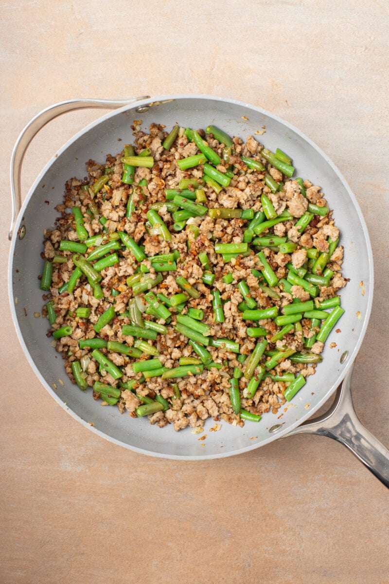 Green beans added to ground pork in a saute pan to make a stir fry.