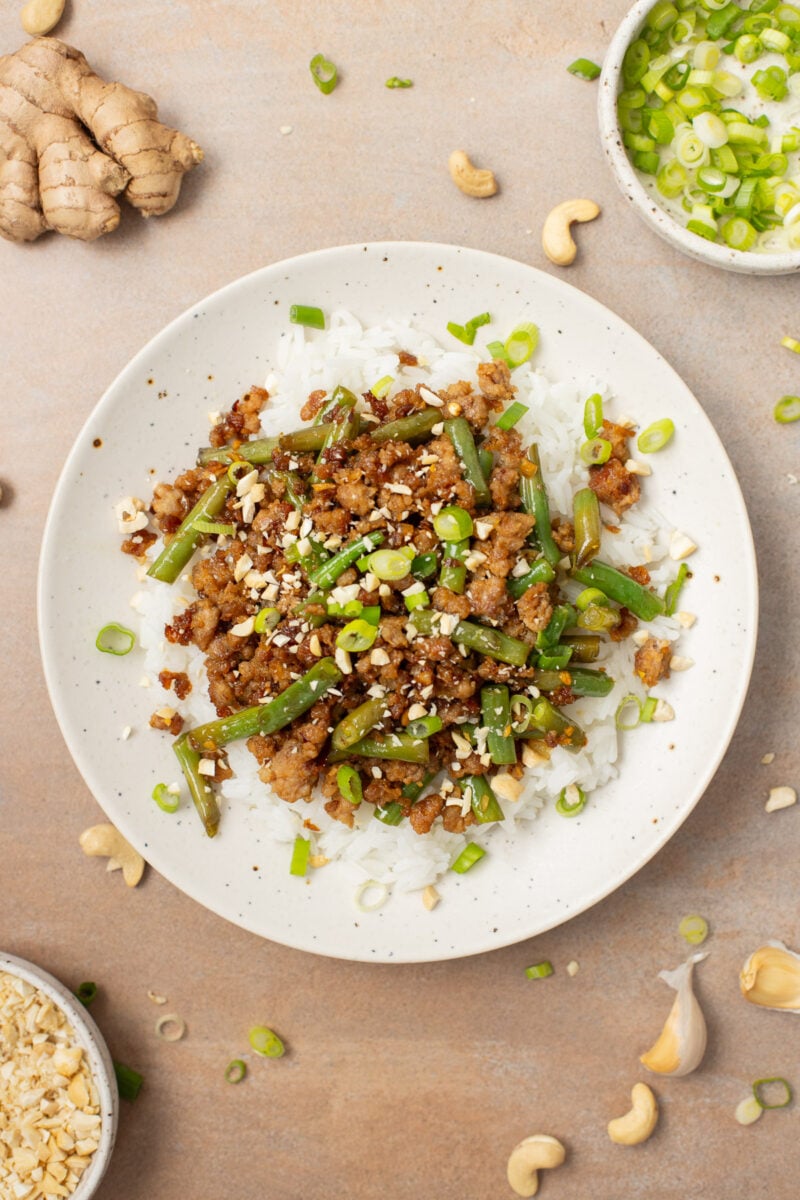 Healthy ground pork stir fry on a bed of white rice on a plate with ginger and green onions on the side.