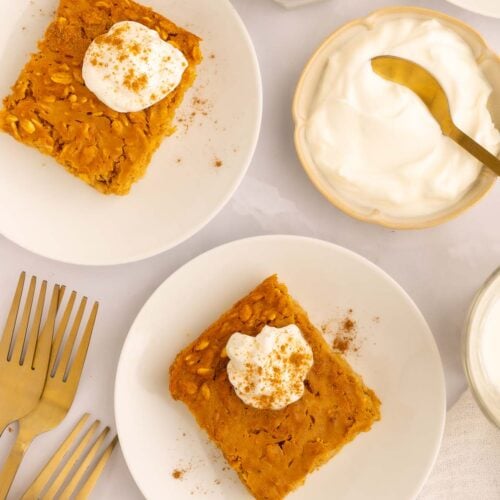 Pumpkin Baked Oatmeal servings on two white plates topped with whipped cream and cinnamon with a small bowl of whipped cream on the side.