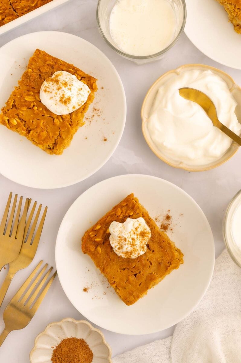 Pumpkin Baked Oatmeal servings on two white plates topped with whipped cream and cinnamon with a small bowl of whipped cream on the side.