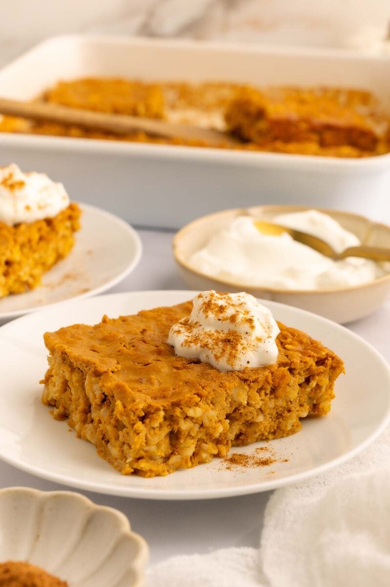 A serving of pumpkin baked oats on a white plate with a dollop of greek yogurt on top with the baking dish in the background.