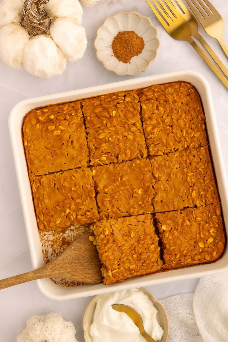 Pumpkin baked oatmeal in a baking dish after being cut into squares with one serving taken out and a wooden spoon scooping out another piece.
