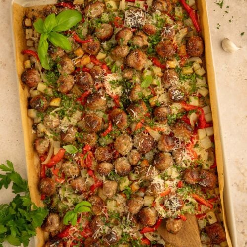 Sausage, peppers, potatoes, and onions on a sheet pan with a wooden spoon in it.
