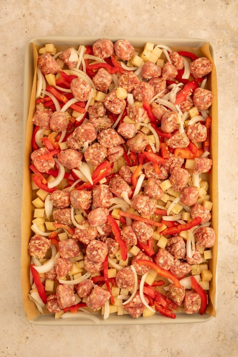 Uncooked Italian sausage pieces on peppers, potatoes, and onions on a sheet pan. 