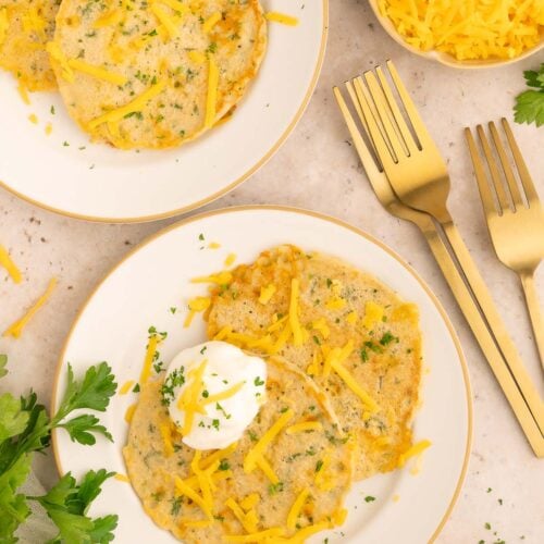 Savory pancakes on a white plate topped with cheddar cheese and fresh herbs.