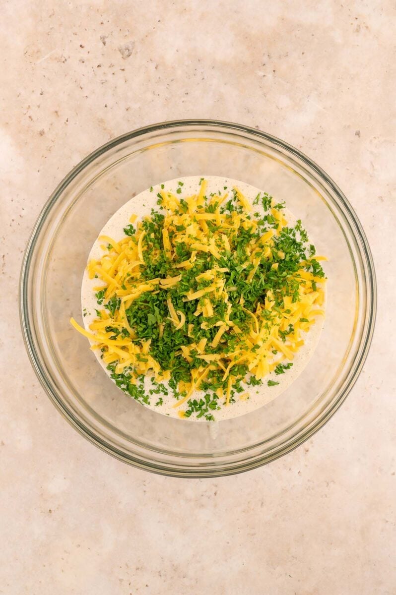 Savory pancake batter in a clear large bowl with shredded cheese and fresh parsley on top before folding in the herbs and cheese.