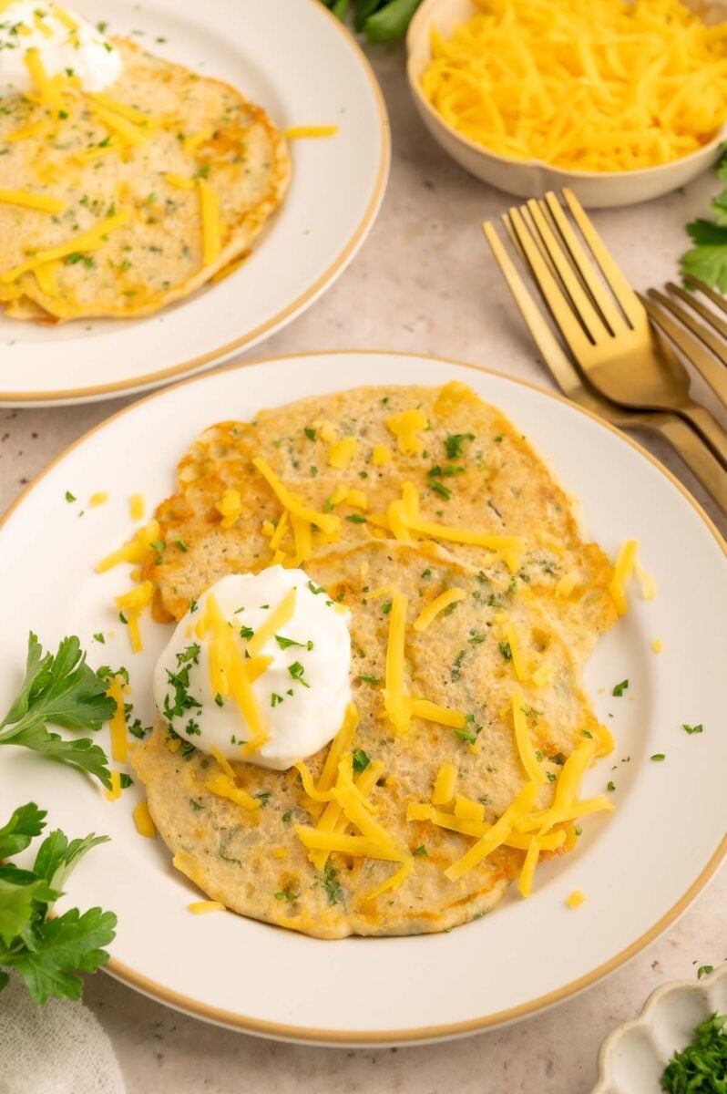 Two savory cheddar pancakes on a white plate topped with a dollop of sour cream and scattered with shredded cheese and fresh herbs. 