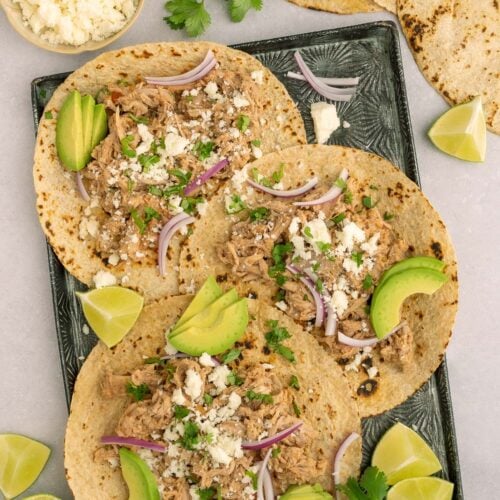 Three pulled pork tacos on corn tortillas garnished with cotija cheese, red onions, and sliced avocado on a black sheet pan with more ingredients on the side.