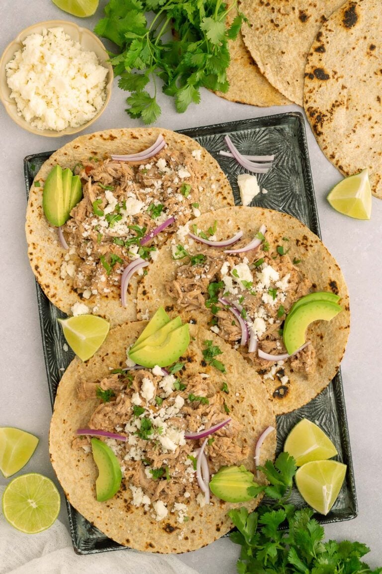 Three pulled pork tacos on corn tortillas garnished with cotija cheese, red onions, and sliced avocado on a black sheet pan with more ingredients on the side.