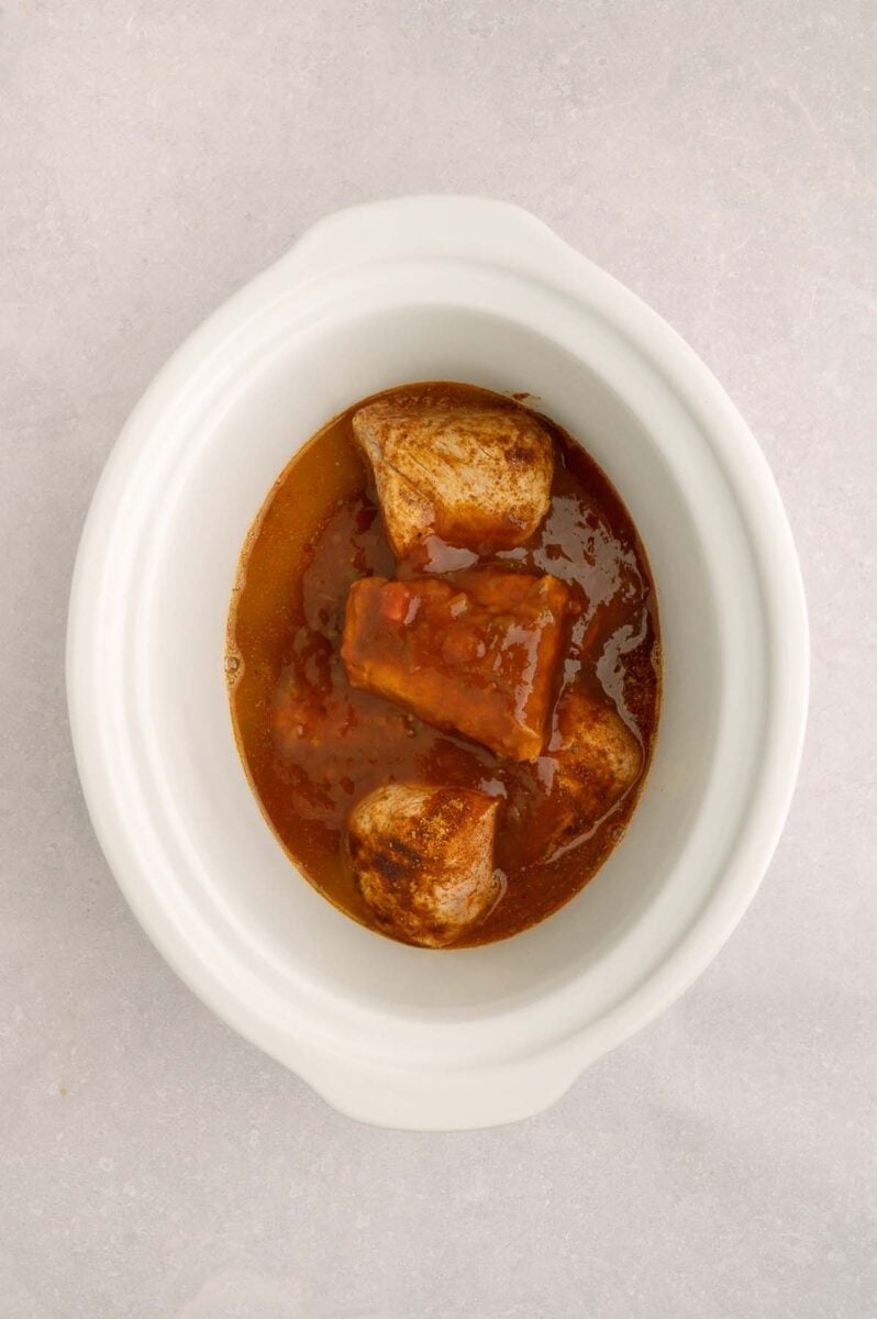 Pork tenderloin pieces in a white crockpot base with taco seasoning, lime juice, salsa and chicken stock on top. 