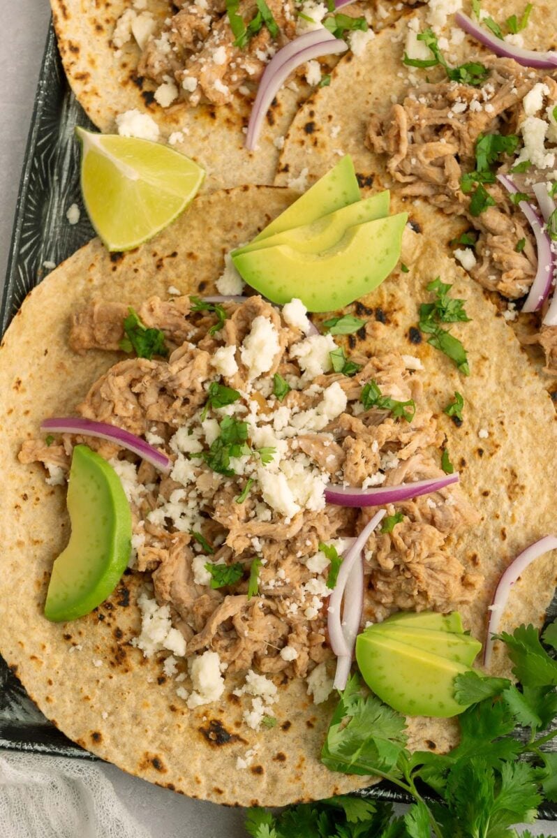 A crockpot pulled pork taco on a black baking sheet garnished with avocado slices, red onions, and cotija cheese.