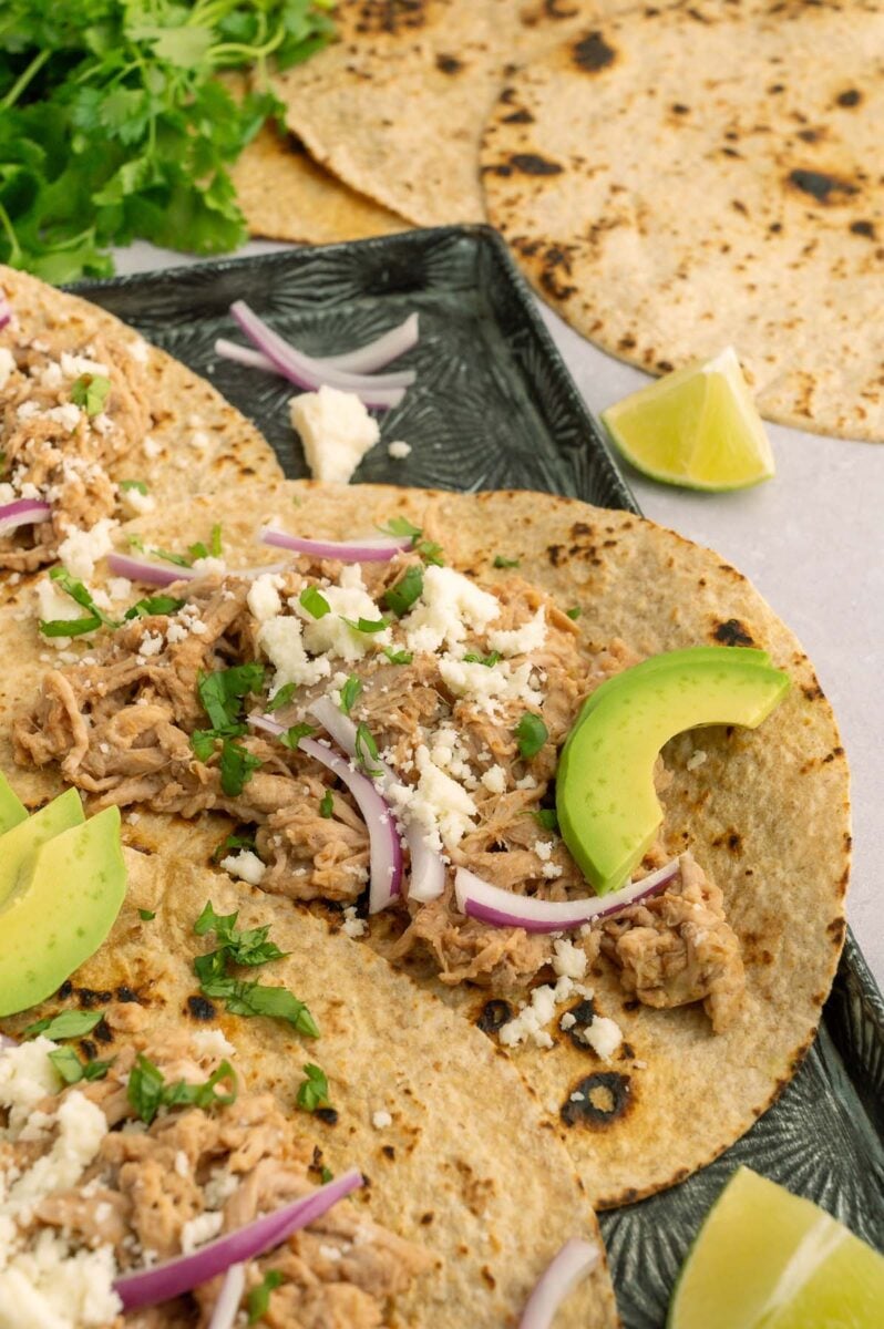 Slow cooker pulled pork tacos on a black sheet pan garnished with onions, avocado, and cheese.