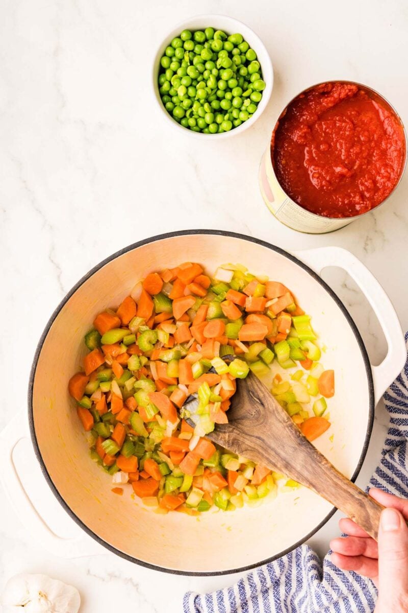 Carrots, celery, and onions cooking in a dutch oven with a wooden spoon in it and a bowl of peas and can of tomatoes on the side.