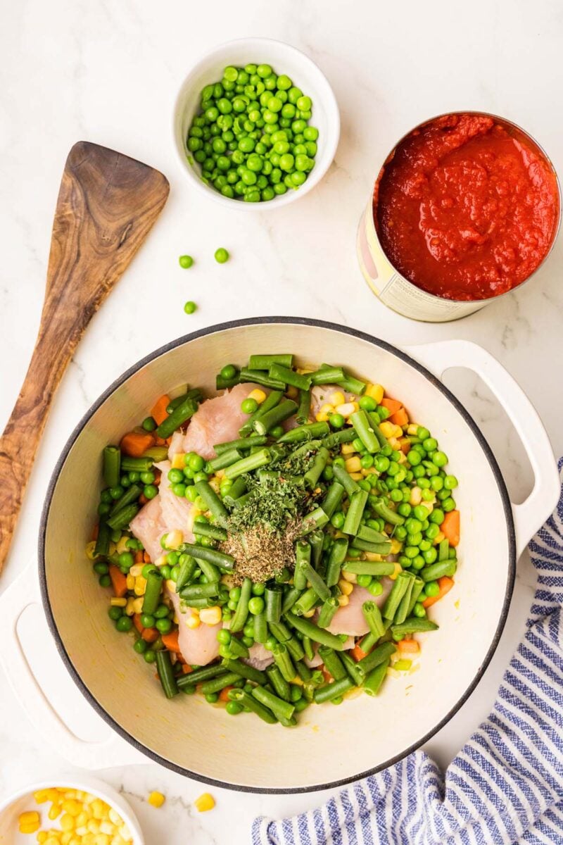 Uncooked chicken breast and veggies in a dutch oven with a bowl of peas and can of tomatoes on the side. 