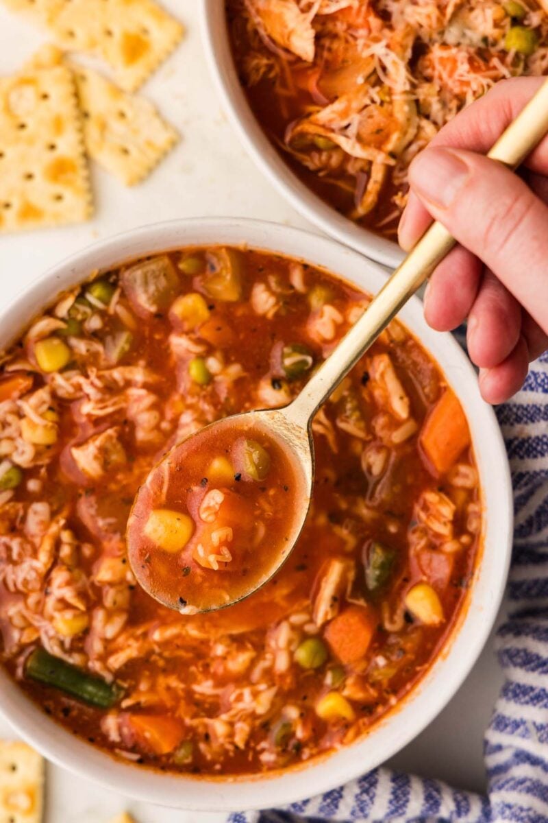 A spoon getting a spoonful of alphabet soup out of a bowl with another soup bowl and crackers on the side.