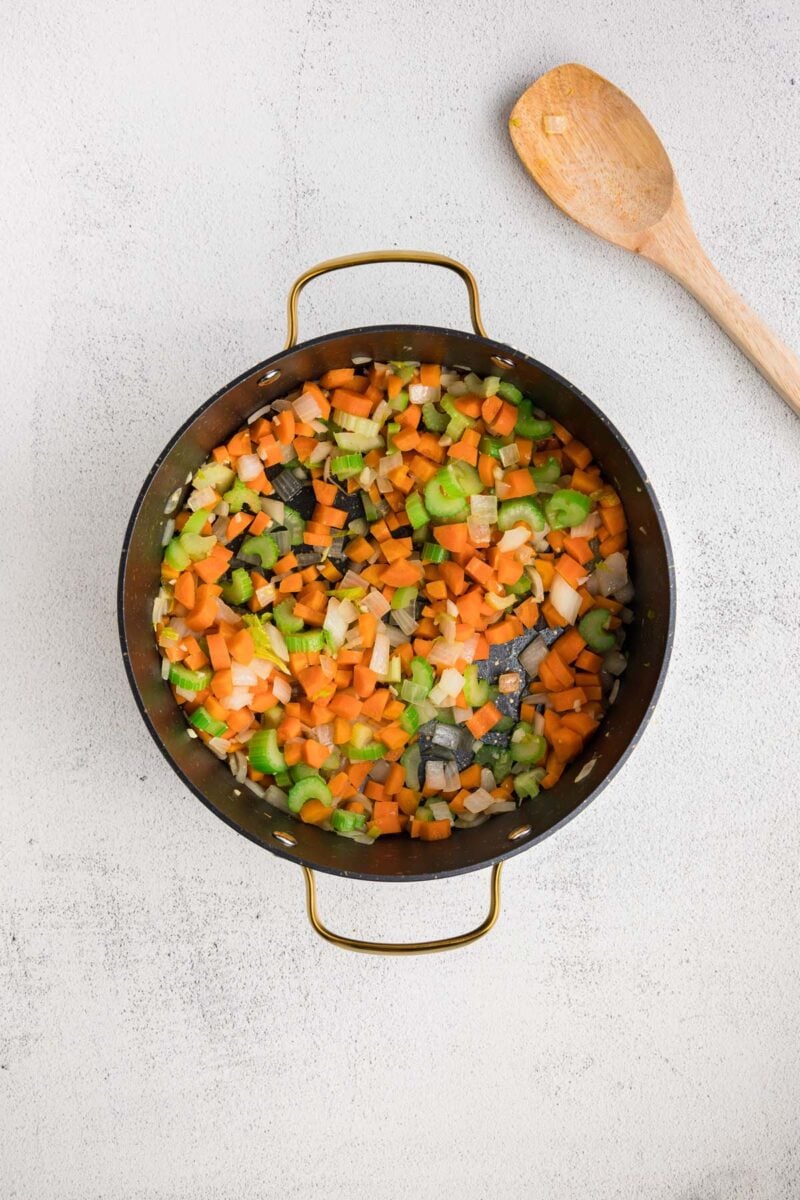 Carrot, celery, onion and garlic cooked in a large pot with a wooden spoon on the side.