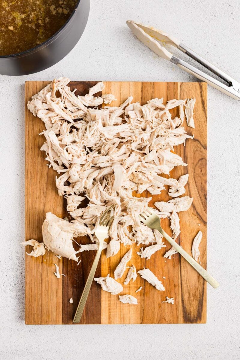 Chicken shredded on a wooden cutting board with two forks in it with a pot of soup and tongs on the side. 