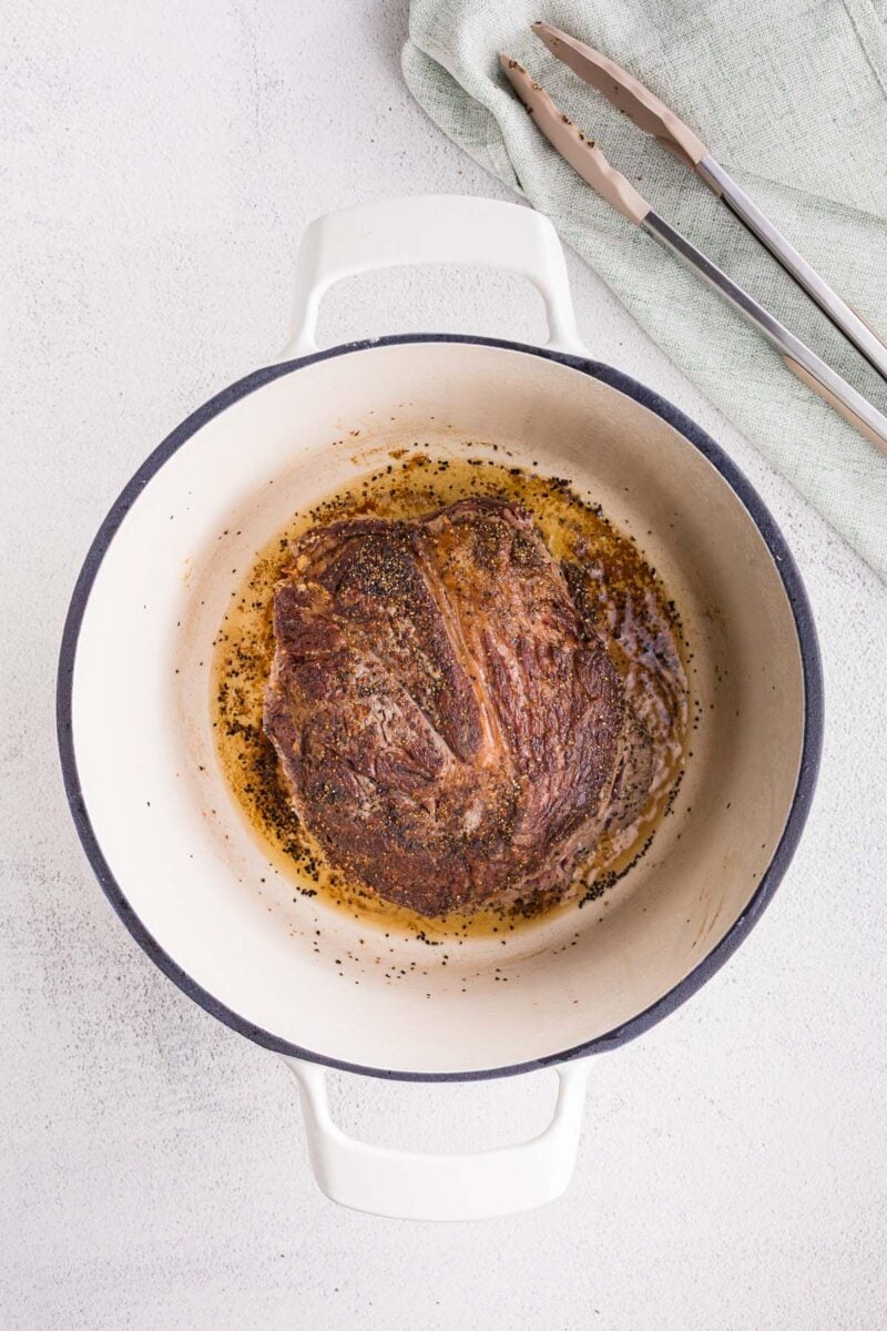 A chuck roast browned in a dutch oven with tongs on the side.