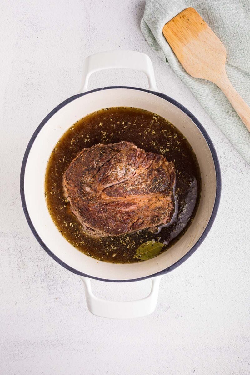 A seared chuck roast in a dutch oven with broth and spices around it before cooking with a wooden spoon on the side.