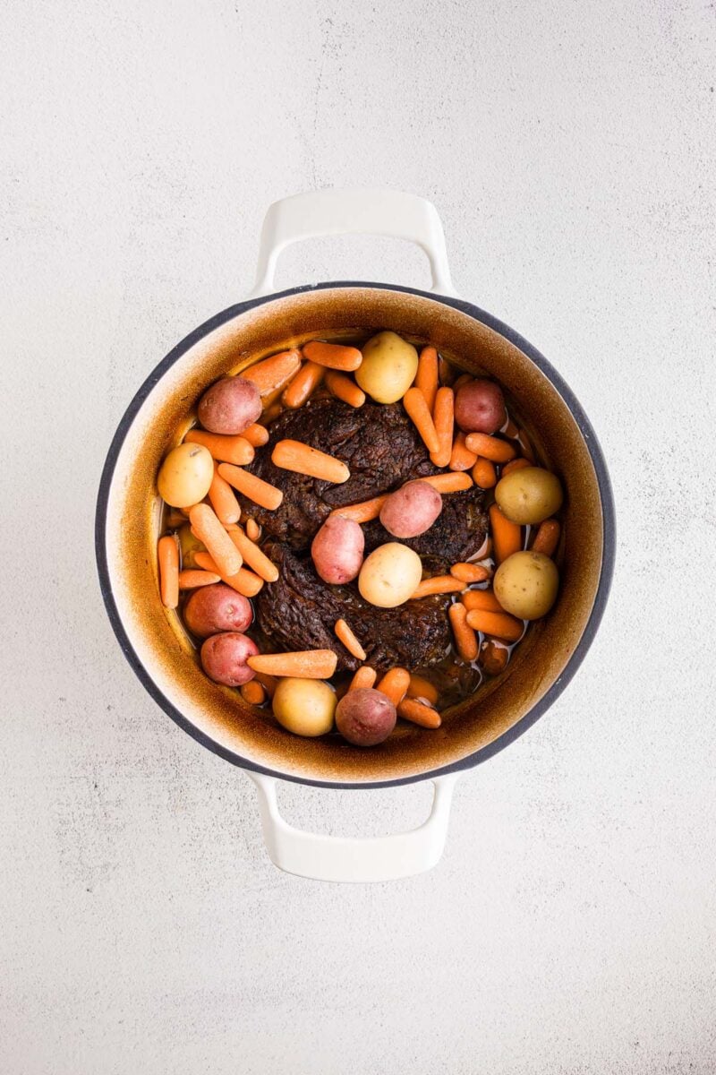 Uncooked carrots and baby potatoes added on a partially cooked pot roast in a dutch oven. 