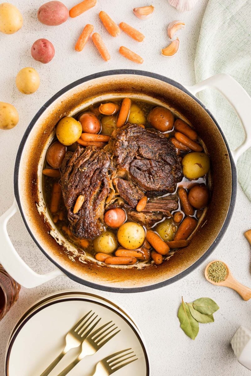 Pot roast, carrots, and potatoes in a dutch oven with more ingredients, plates and forks on the side.
