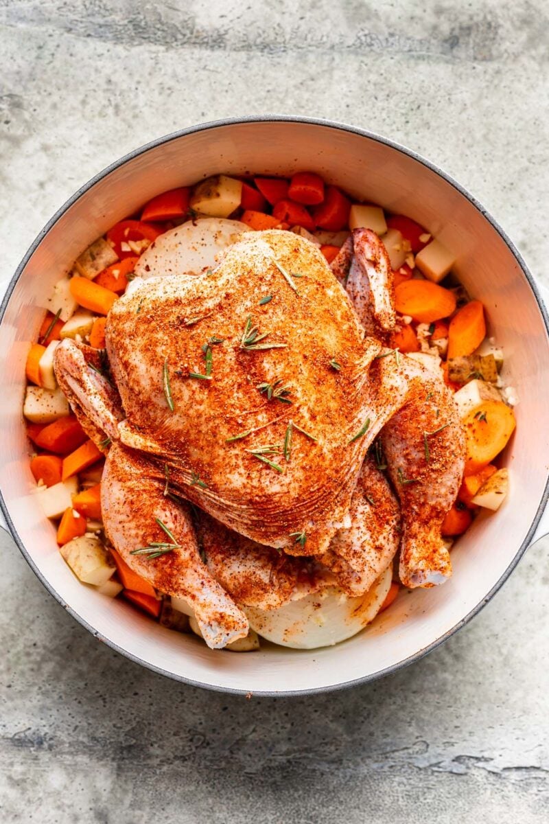 Uncooked whole seasoned chicken on top of vegetables and potatoes in a large dutch oven.
