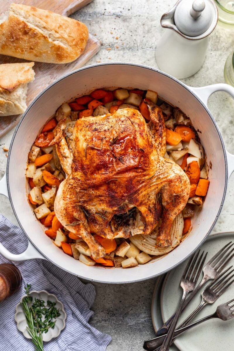 A whole chicken on top of veggies and potatoes in a dutch oven with a french loaf of bread on the side.