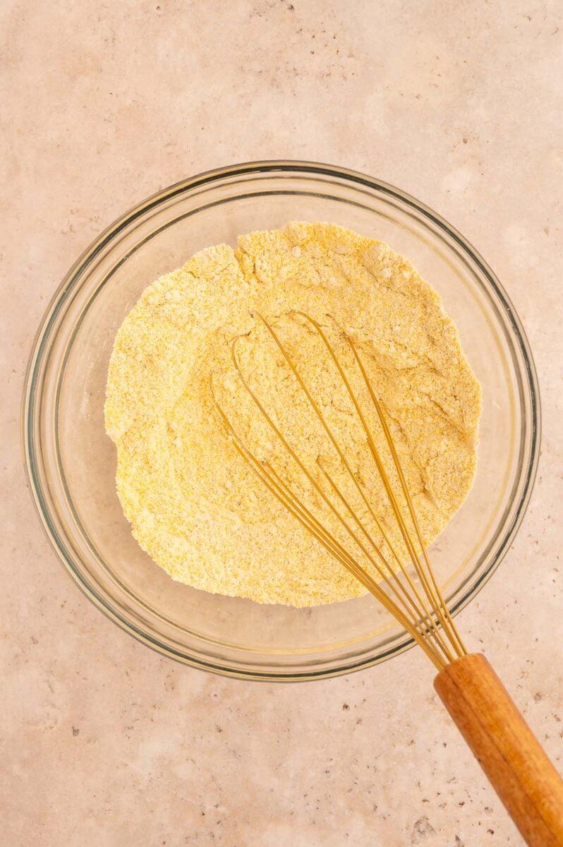 Dry ingredients for healthy cornbread in a clear glass bowl with a whisk in it.