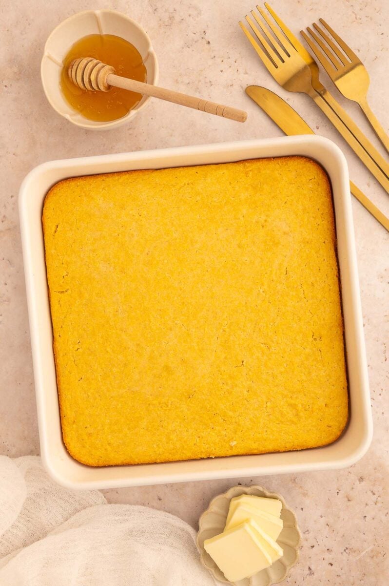 Healthy cornbread in a pan with a bowl of honey with a honey dipper on the side.