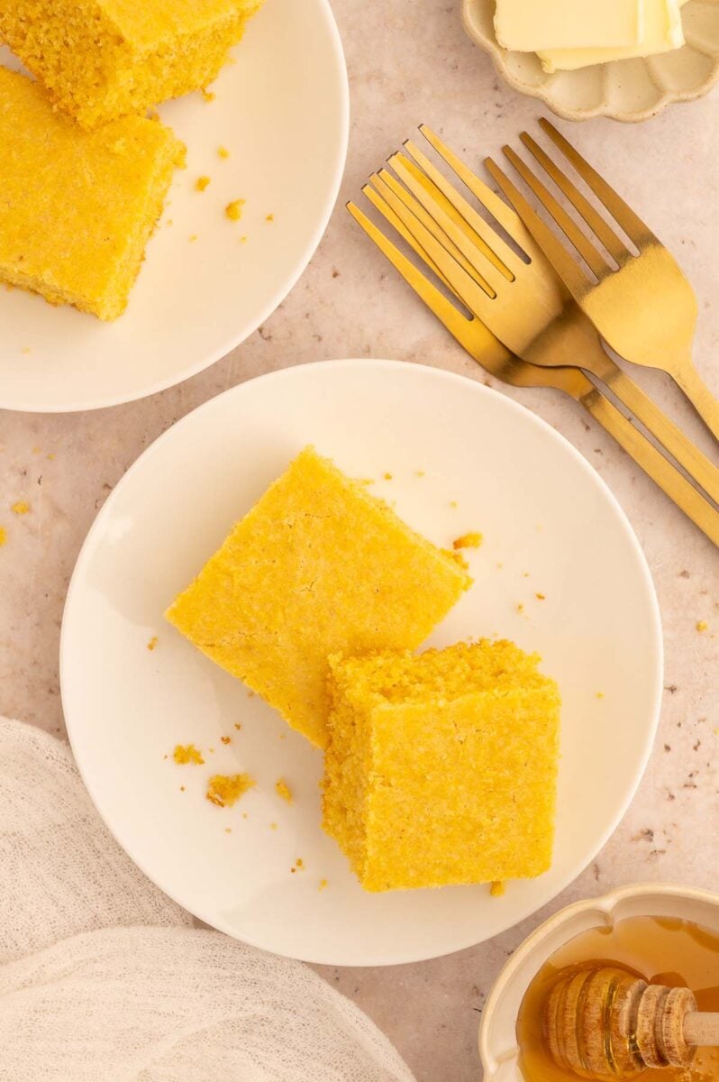 Two pieces of healthy cornbread leaning on each other on a white plate with another plate of corn bread on the side.