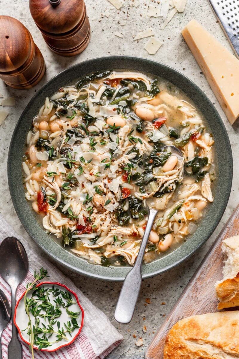 Tuscan Chicken Soup in a grey bowl with a spoon in it and parmesan cheese rind on the side.