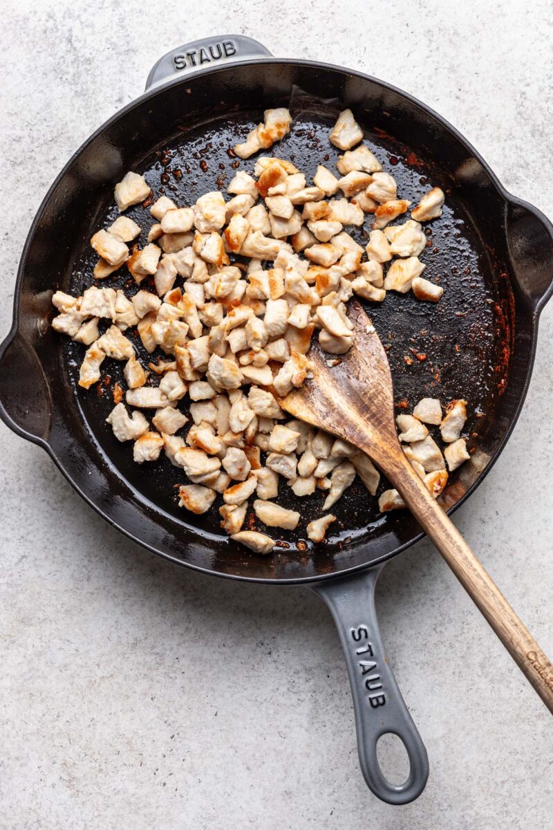 Browned chicken pieces in a cast iron skillet with a wooden spoon in it.